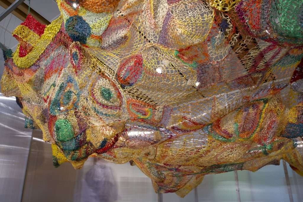 A photo of an artwork made of thin ropes of various colours and hung from the ceiling like a fishing net.