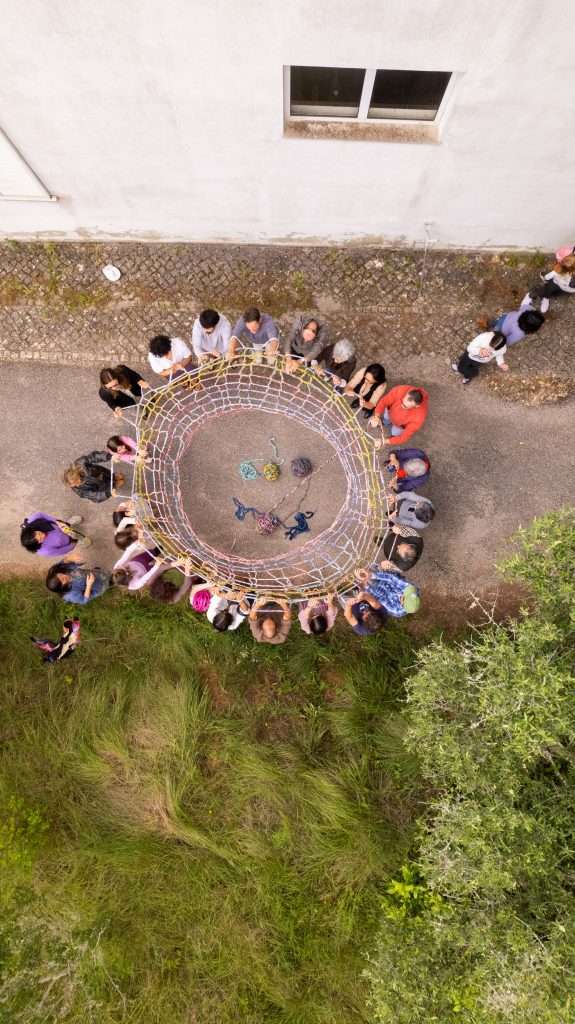 A photograph from above of a group of people taking part in an artistic activity: they all have pieces of a coloured net in their hands and they are standing in a circle